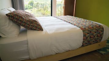 A bed with white linens and a colorful geometric-patterned pillow, set against a green wall with a view of greenery outside.