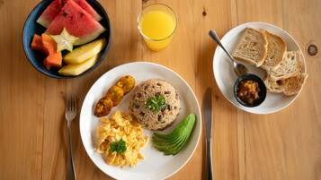 A breakfast setting with a plate of scrambled eggs, a rice ball, fried plantains, and a bowl of fruit.