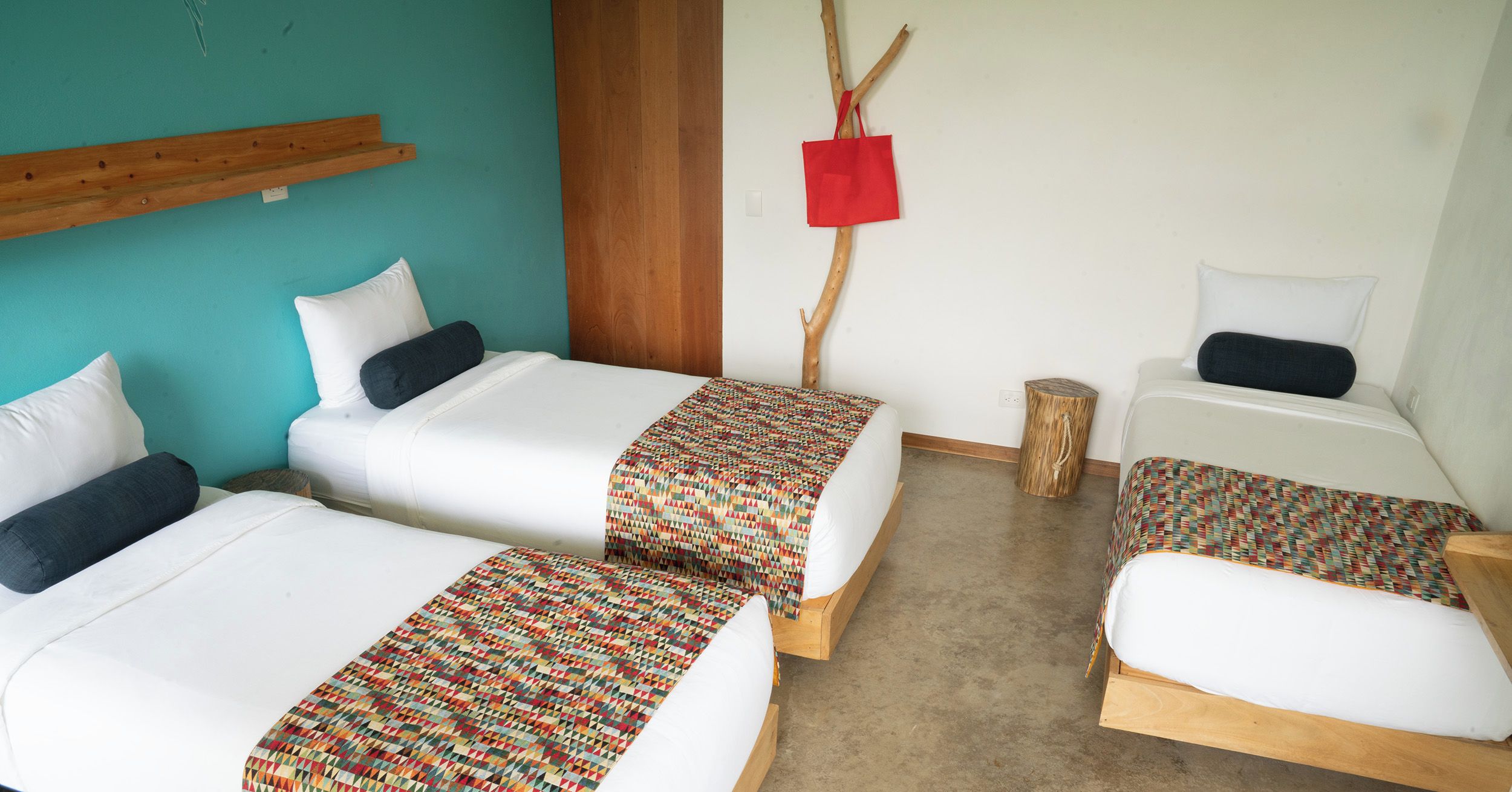 A room with two single beds, a small table, and a red bag hanging on a branch.