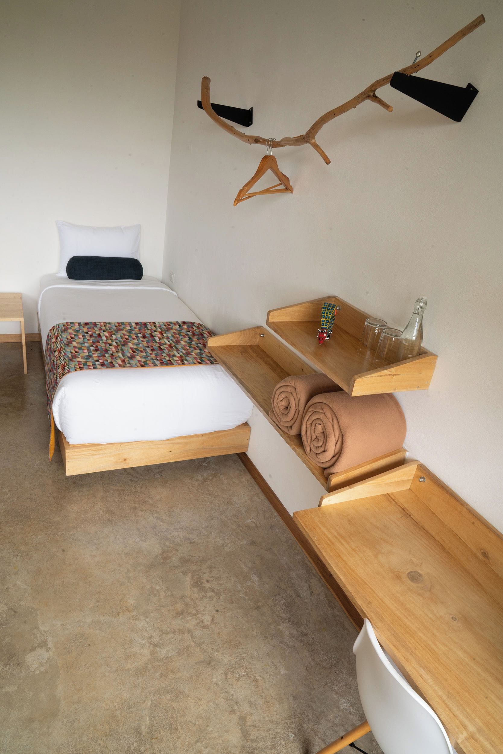 A bedroom with a bed, a wooden desk, a hanging branch, and a small shelf with a bottle and glasses.