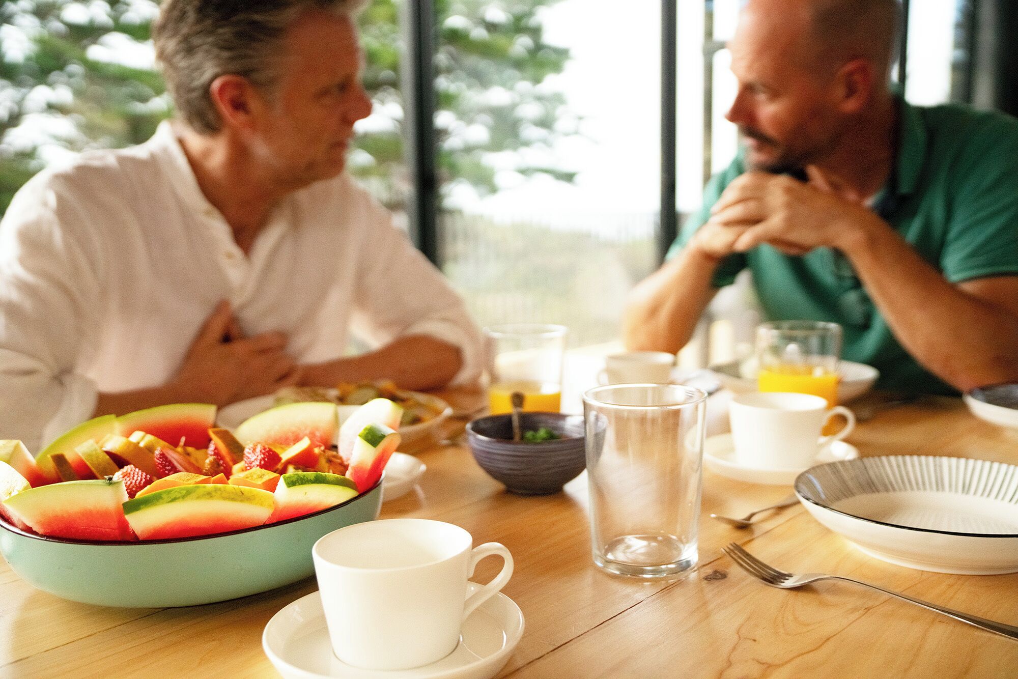 Un desayuno con frutero, tazas y jarra sobre una mesa de madera.