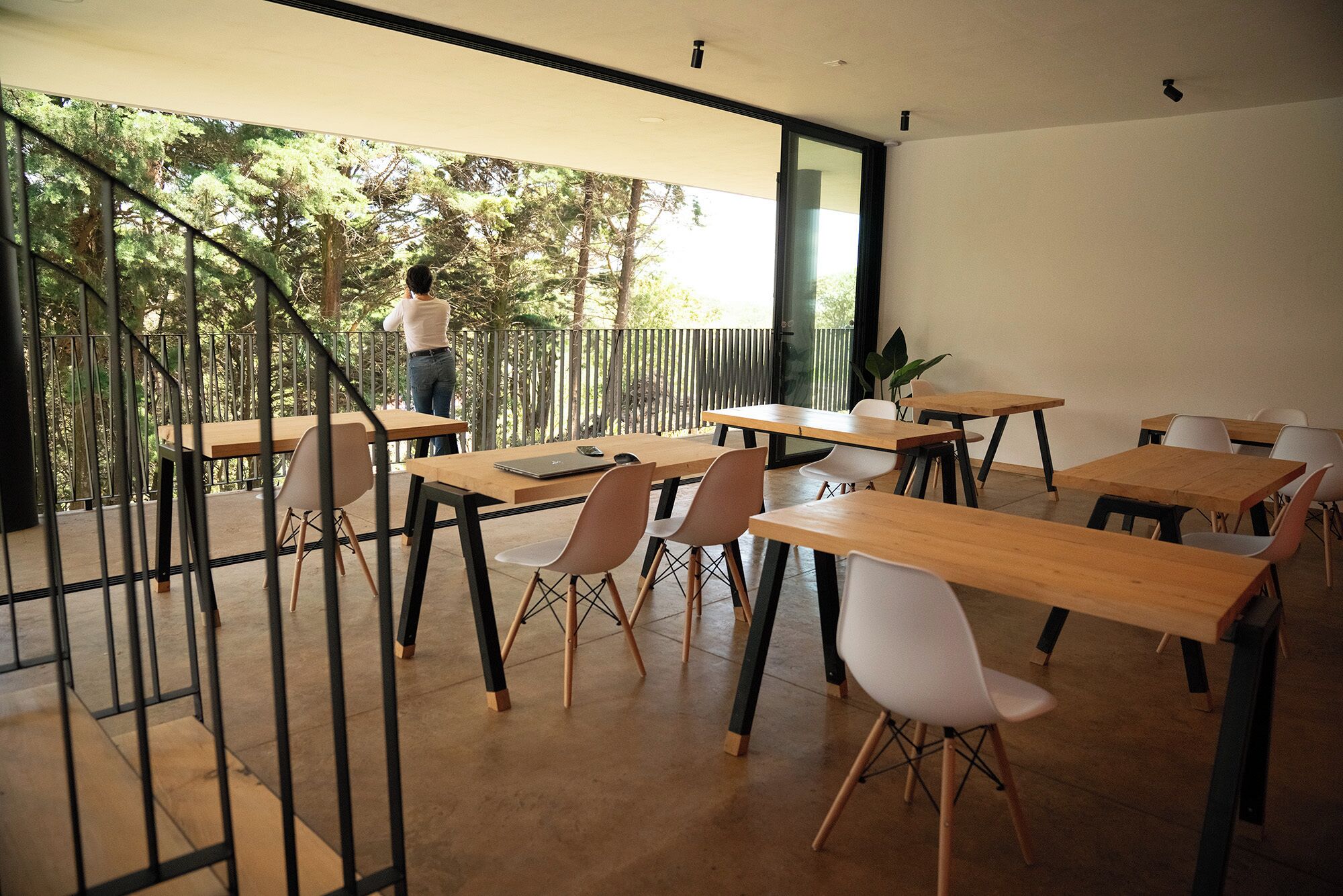 A modern dining area with wooden tables and chairs, a staircase leading to an outdoor area, and a large window providing a view of trees.