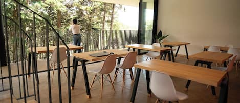 Un comedor moderno con mesas y sillas de madera, una escalera que conduce a un espacio exterior, y un ventanal con vistas a árboles.