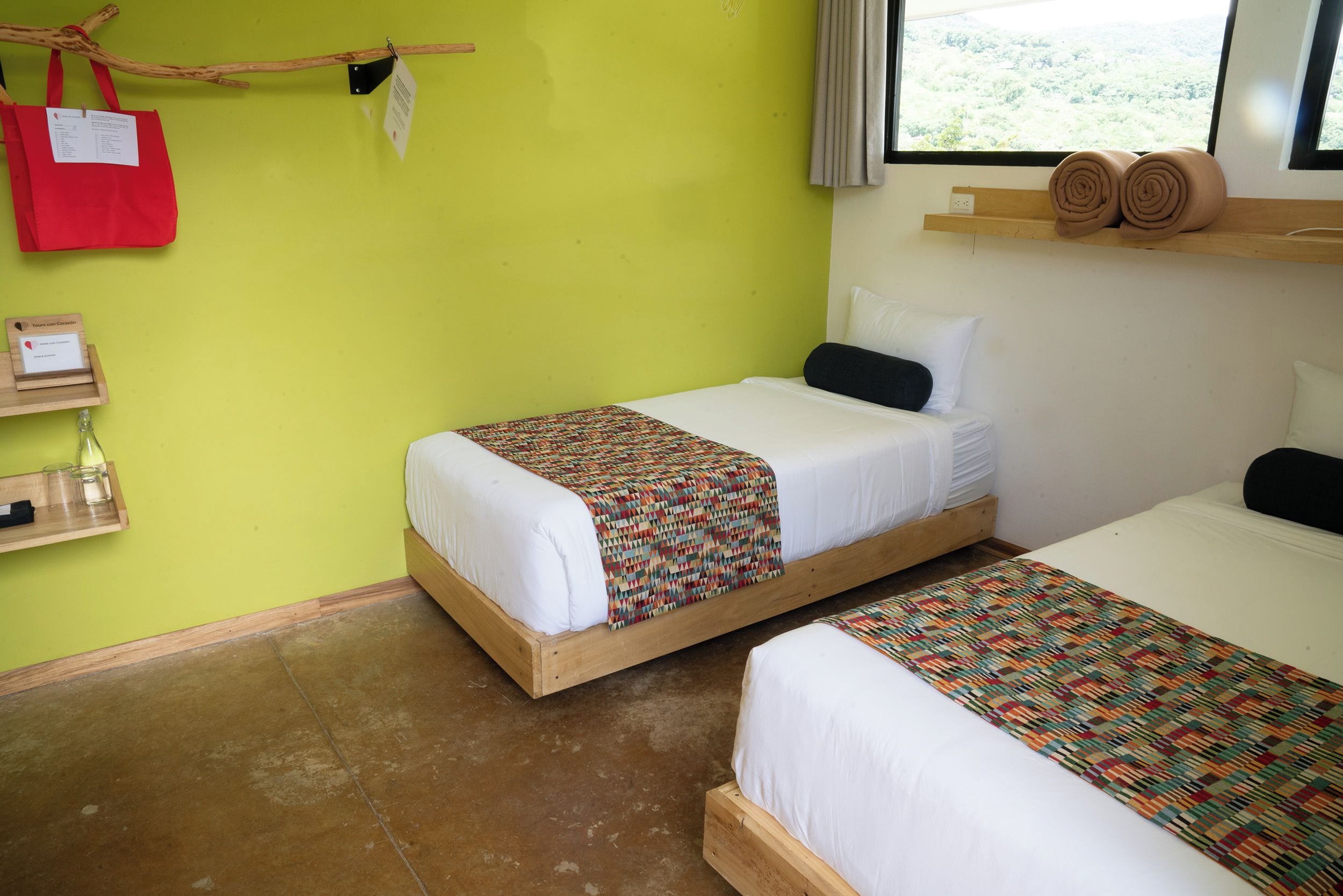 A room with two beds, a red tote bag, a wooden shelf with rolled towels, a small wooden table with glasses and a bottle, and a window with a view.
