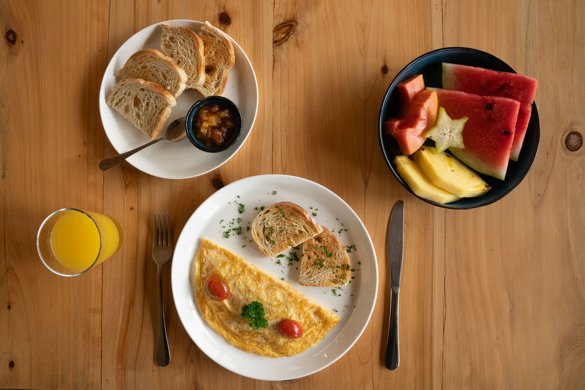 A breakfast setting with toast, a bowl of fruit, a glass of orange juice, and an omelette on a wooden table.