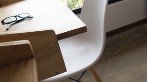 Une table en bois sur laquelle reposent des lunettes, une chaise blanche et une vue sur un balcon doté d’une balustrade et bordé de verdure.