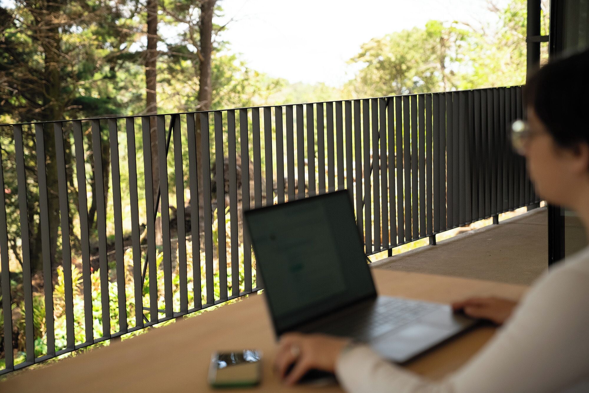 A person using a laptop at a table with a view of trees through a fence.