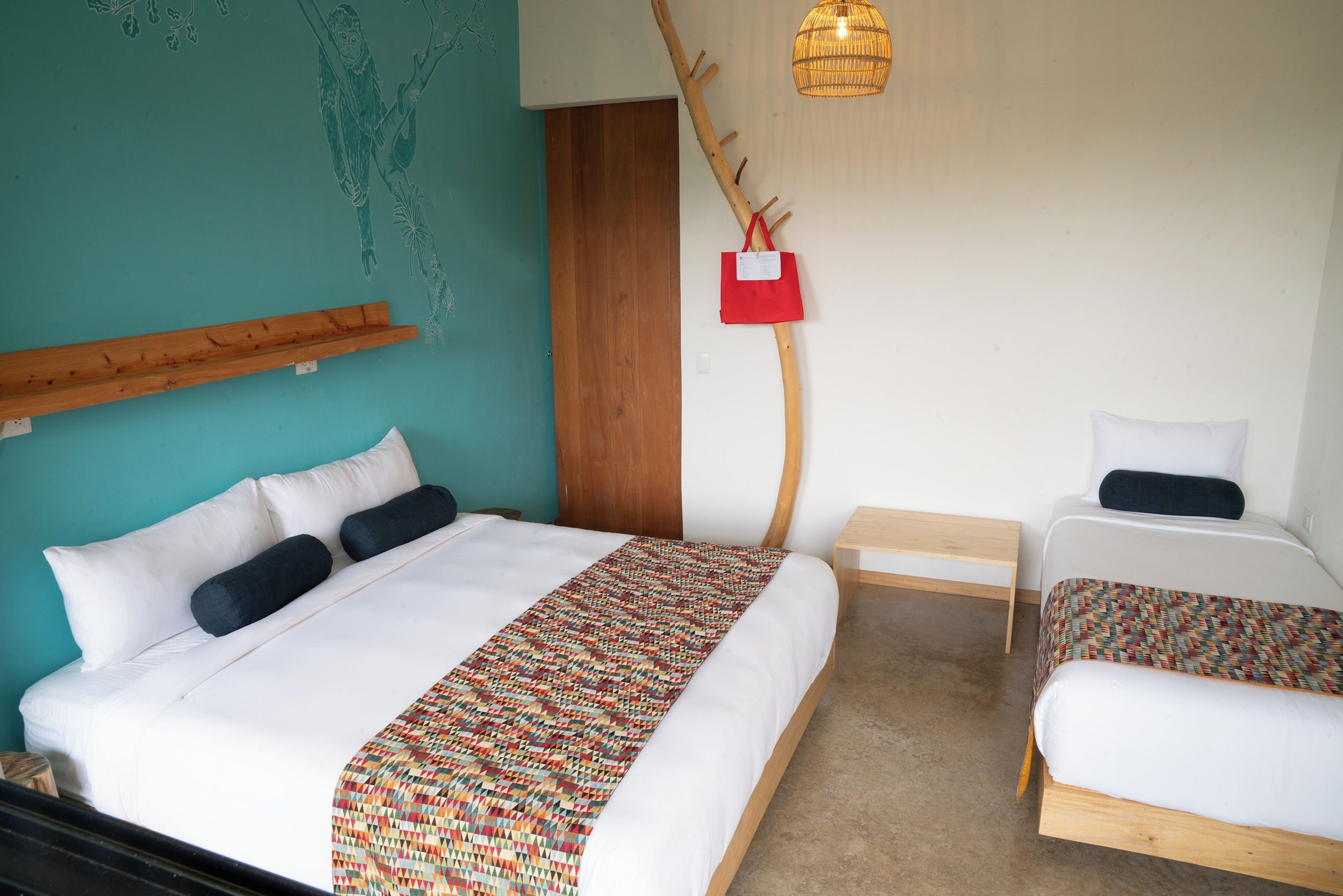 A hotel room with a bed, a smaller bed, a wooden headboard, a wall art piece, a hanging light fixture, and a small table.