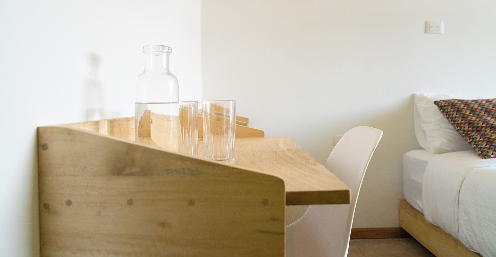 A wooden table with a bottle and two glasses, a white chair, and a bed with a colorful pillow in a simple room.