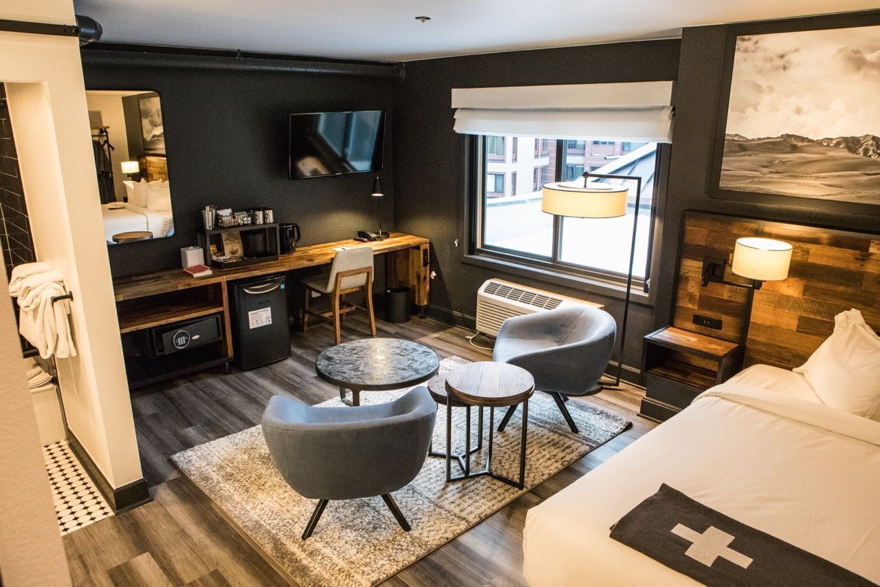 A hotel room with a bed, a desk with a chair, a small round table, two armchairs, a TV, and a window with a view of buildings.