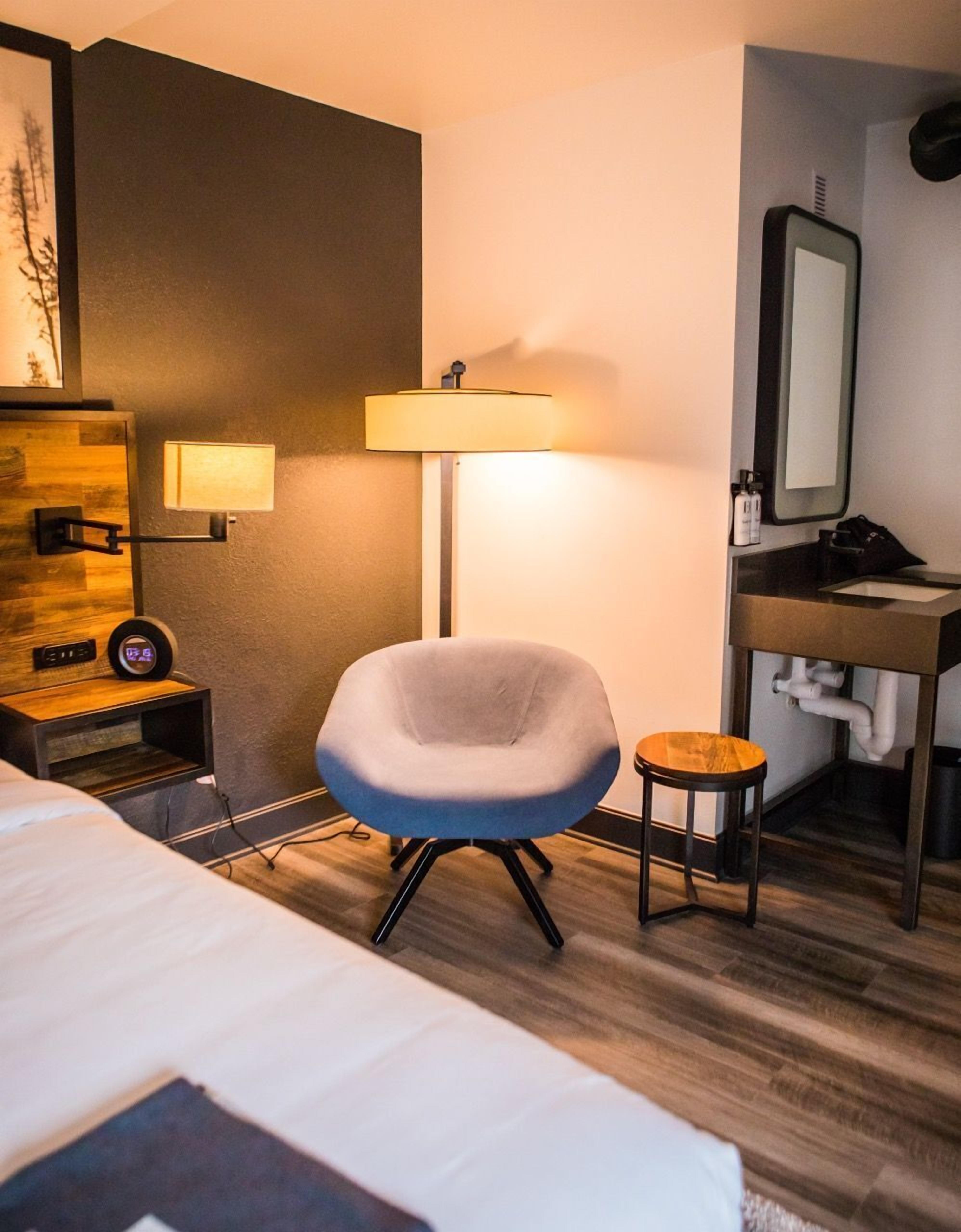 A hotel room with a bed, a grey armchair, a small stool, a bedside table with a lamp, a mirror, and a wooden accent wall.