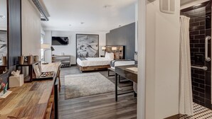 Moderni hotellihuone, jossa on puulattia, suuri sänky, työpöytä tuolilla, kylpyhuone suihkulla sekä suuri taulu seinällä.