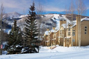 Un edificio cubierto de nieve con una escalera que conduce a la entrada, rodeado de árboles y montañas al fondo.