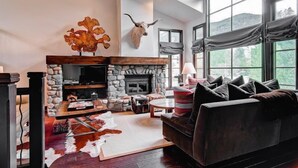 Una sala de estar acogedora con chimenea de piedra, una cabeza de animal montada y grandes ventanales que ofrecen vistas al exterior.