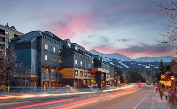 A multi-story hotel with a modern design, featuring a mix of wood and metal facades, located in a mountainous area with a view of snow-capped peaks.