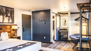 A modern hotel room with a large bed, a dark door, a mirror, a seating area with a chair, and a wooden bench.
