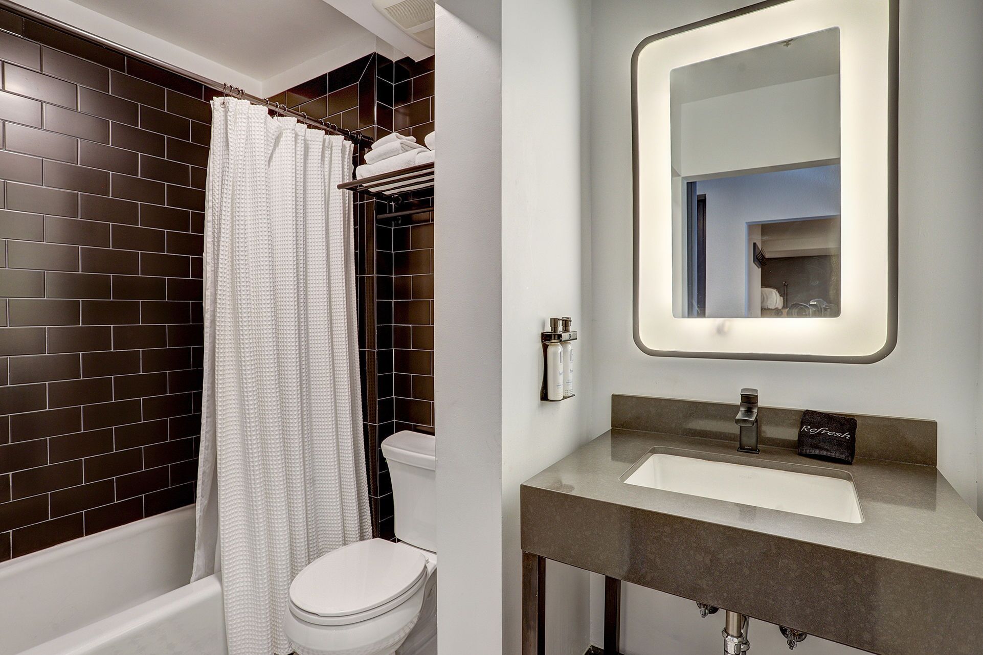 A bathroom with a white toilet, a shower with a white curtain, a mirror with a light, and a sink with a faucet.