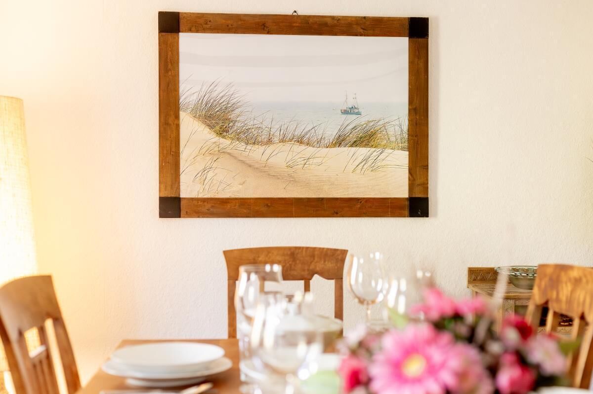 A dining area with a table set for two, a framed picture of a beach scene on the wall, and a vase of flowers on the table.