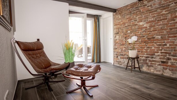 A modern living room with a leather armchair and ottoman, a brick wall, and a view of the outdoors. - Anno 1895 - Holiday home for up to 4 people, pets welcome (Manslagt)