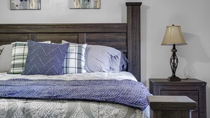 Ein ordentlich bezogenes Bett mit gemusterten Bettwäsche und Kissen, eine Nachttischlampe und zwei Blumenbilder an der Wand.