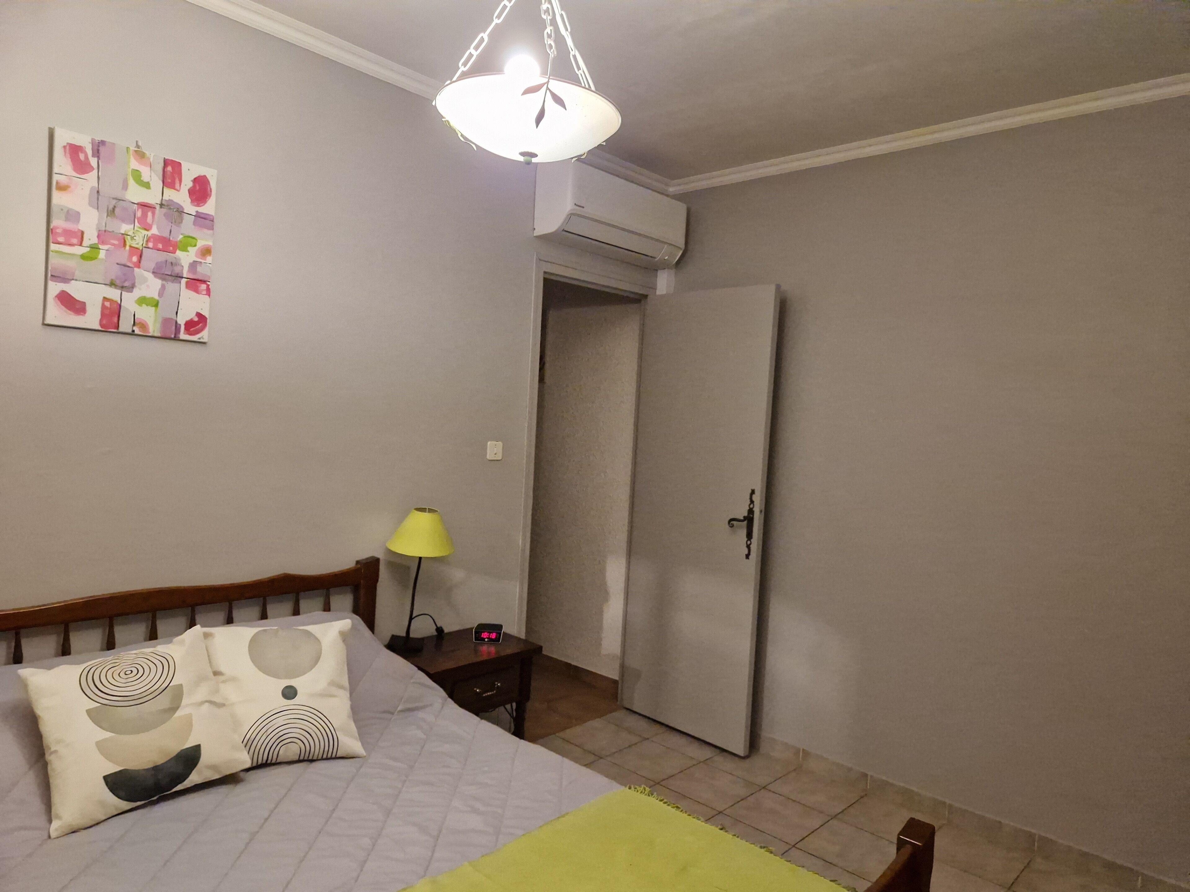 Een slaapkamer met een bed, een nachtkastje met een lamp, een wandklok en een deur.