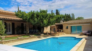 A swimming pool with a stone deck, surrounded by a patio area with outdoor furniture and a view of a house with a tiled roof.