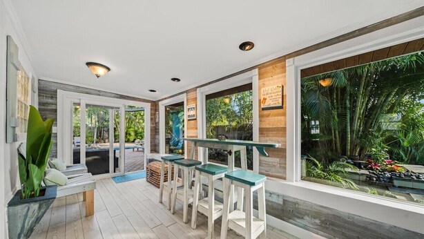 Un'area bar moderna con pavimento in legno, sgabelli bianchi e ampie finestre con vista su un giardino tropicale.