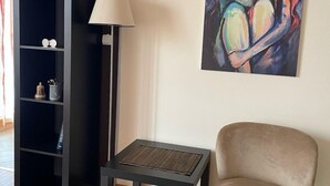 A living room with a beige armchair, a black floor lamp, a black shelving unit, and a colorful abstract painting on the wall. - Couple Studio PG (Podgorica)