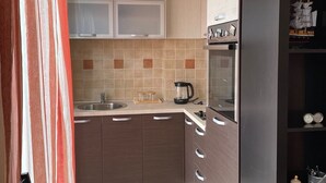 A compact kitchen with a sink, stove, and a coffee maker. There is a dining table with chairs and a window with curtains. - Couple Studio PG (Podgorica)