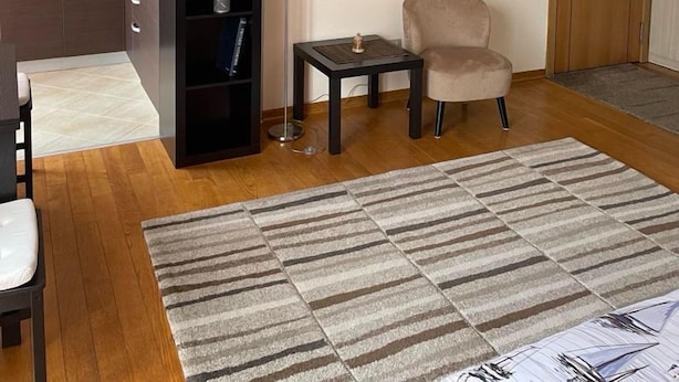 A modern living room with a sofa, a coffee table, and a rug. There is a kitchen area with cabinets and a microwave. A painting is on the wall, and a door is visible in the background. - Couple Studio PG (Podgorica)