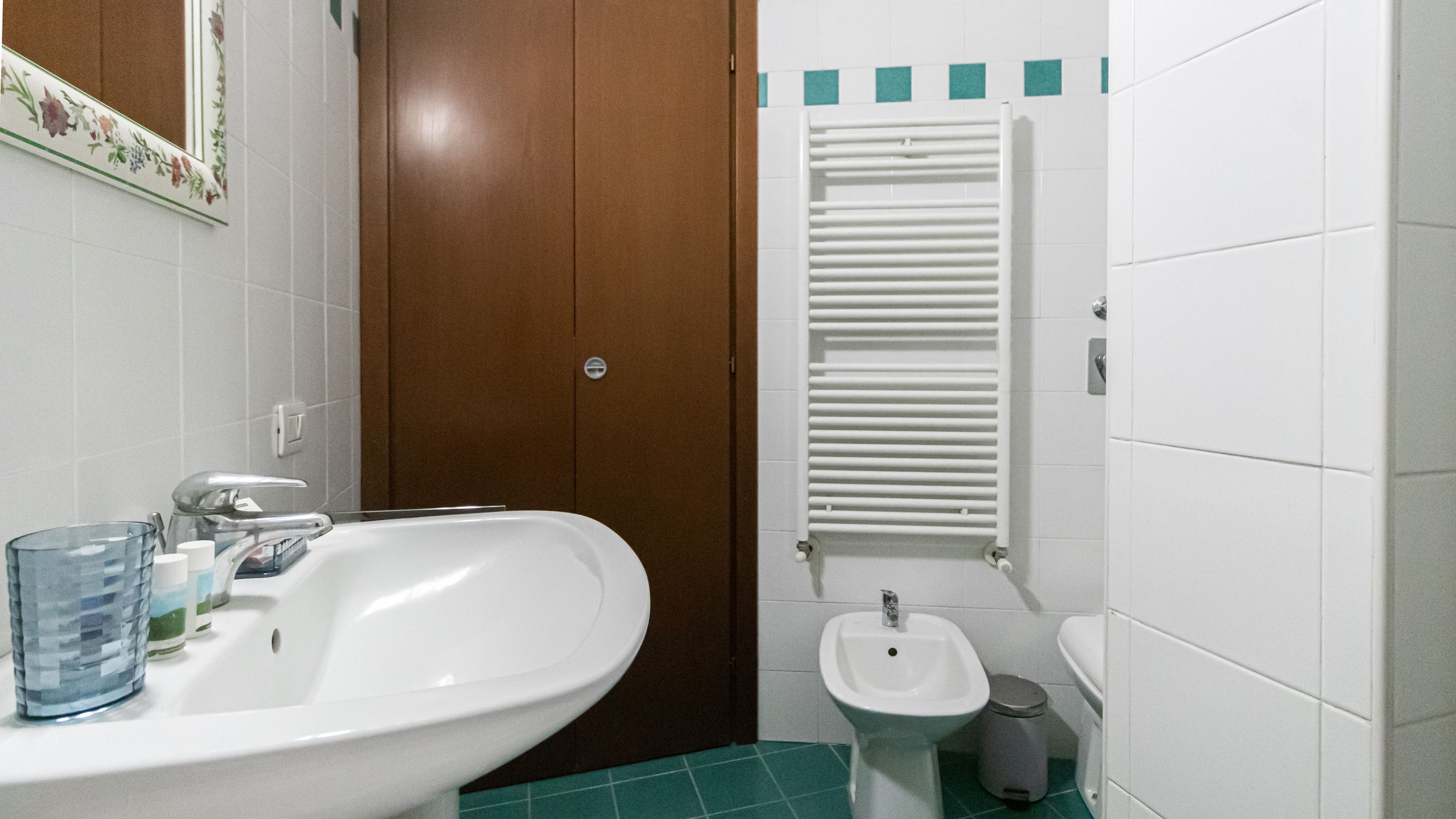 Ein Badezimmer mit einem weißen Waschbecken, einem Spiegel, einer braunen Tür, einem weißen Heizkörper und einem grünen Boden.