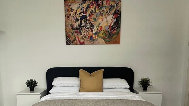 A modern bedroom with a large bed, a tufted ottoman, two bedside tables with plants, and a colorful abstract painting on the wall.
