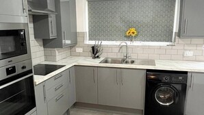 A modern kitchen with grey cabinets, a built-in oven, a dishwasher, and a window with a patterned blind.