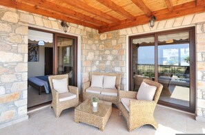 A covered outdoor seating area with wicker furniture, stone walls, and large glass doors leading to a balcony with a view. - Sunset View Villa in Argaka (Argaka)