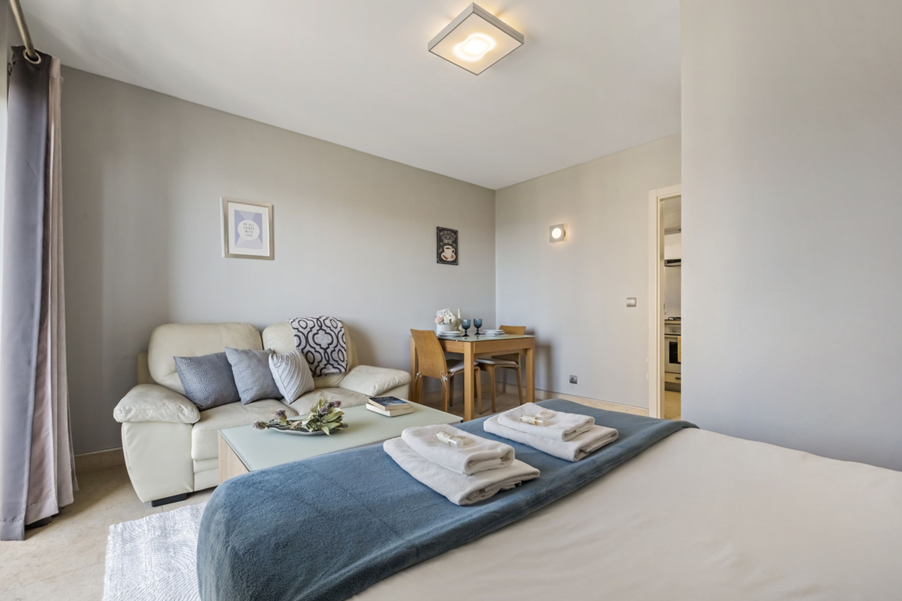 A modern hotel room with a bed, a sofa, a dining table, and a small kitchen area.