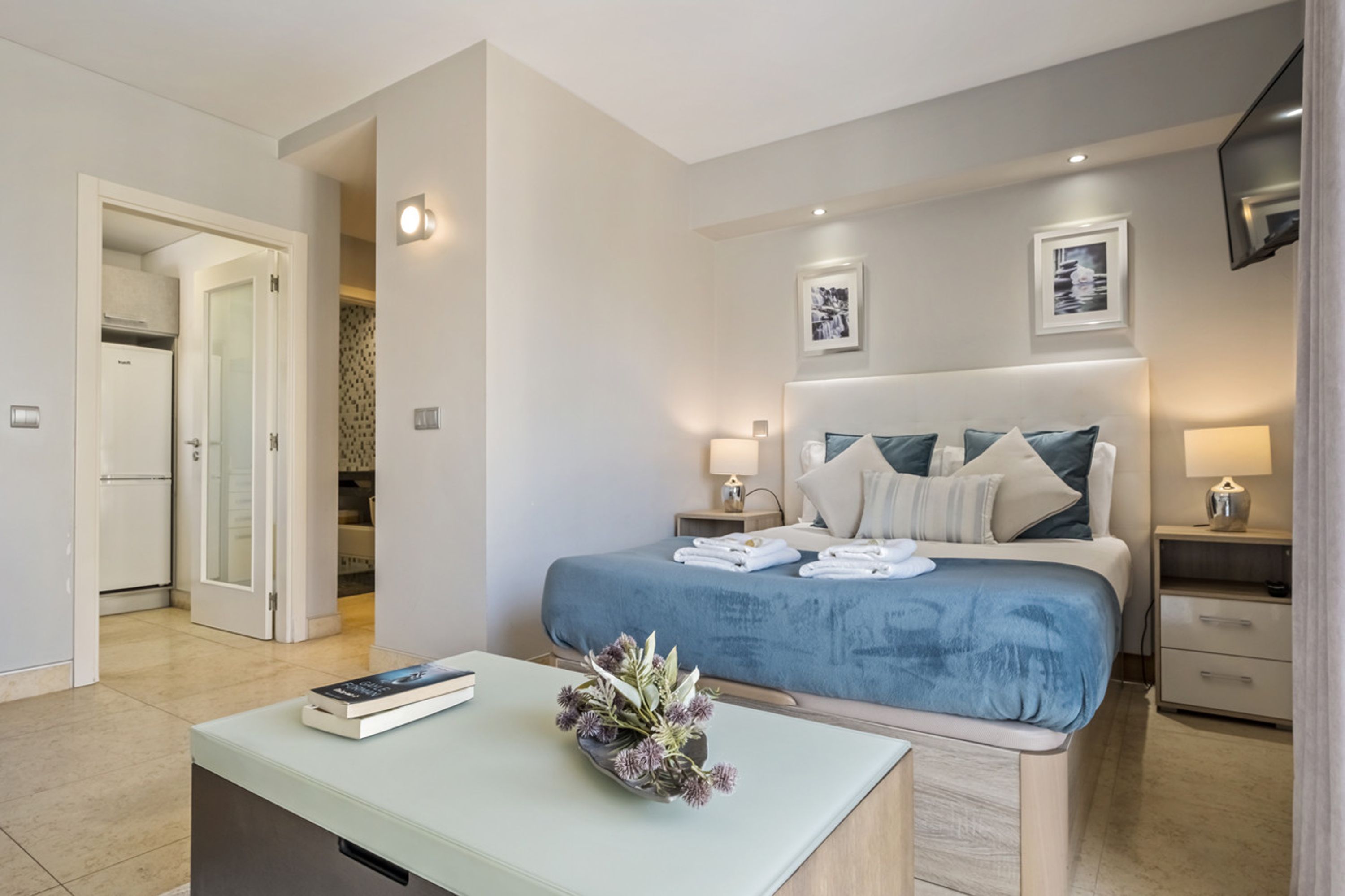 A modern hotel room with a neatly made bed, a bedside table with a book and flowers, and a view of the hallway.