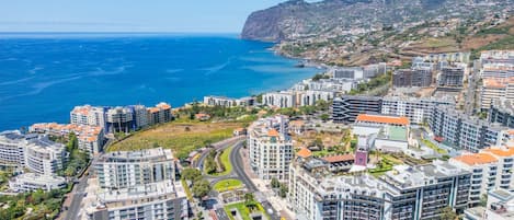A coastal cityscape with modern buildings, a clear blue sea, and a hilly background.