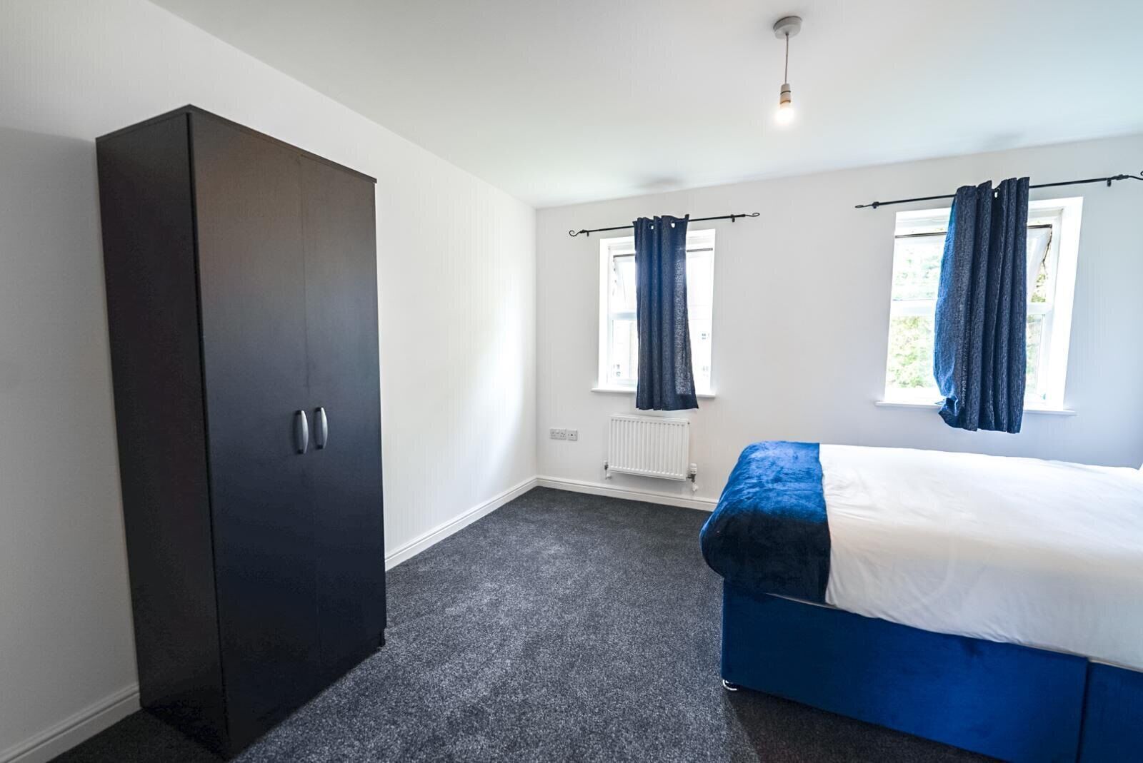 A bedroom with a dark wardrobe, a bed with a blue headboard, two windows with curtains, and a radiator.