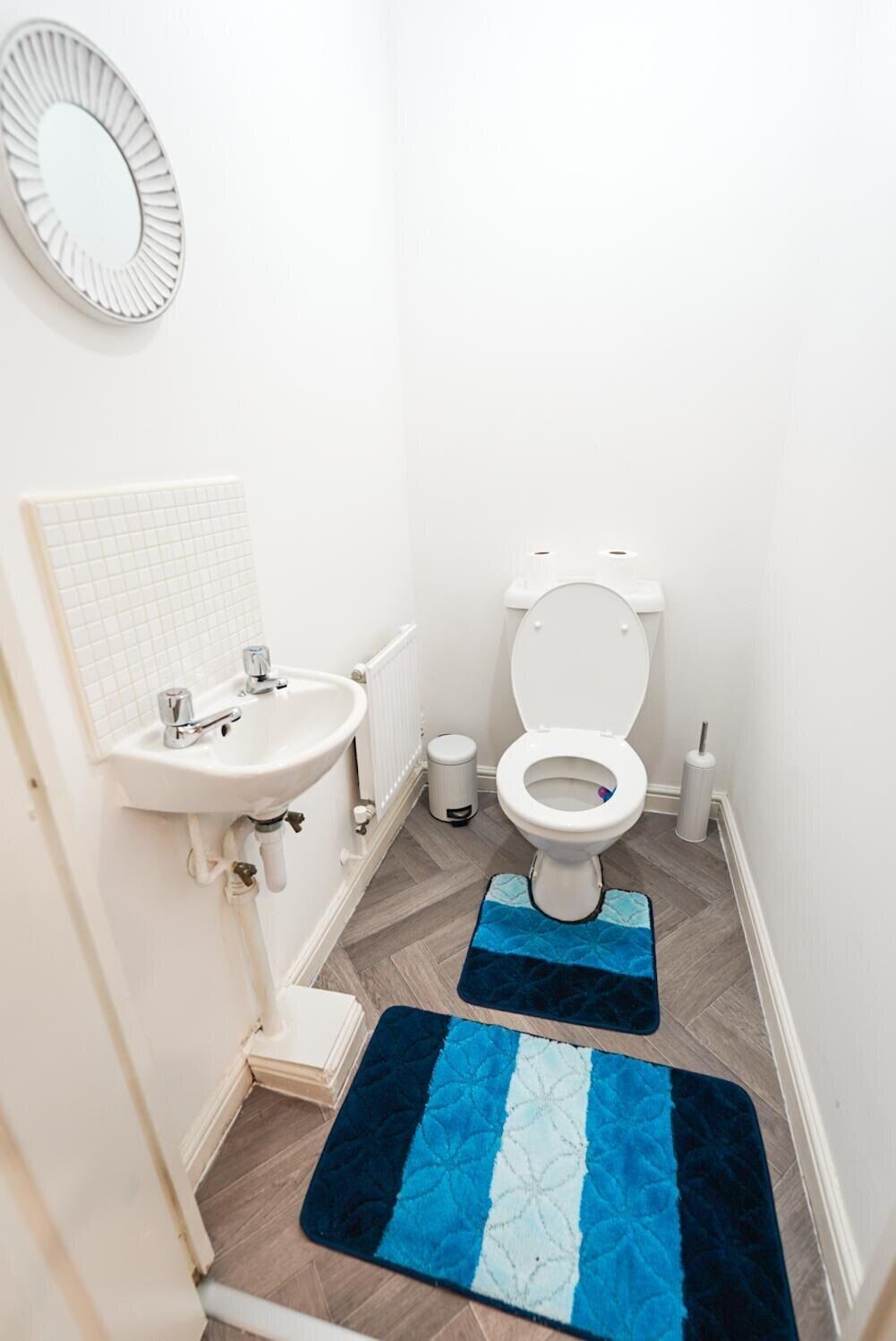 A small, clean bathroom with a white sink, toilet, and a mirror on the wall. There are two blue and white mats on the floor.