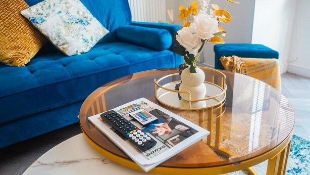 Un salon avec un canapé bleu, une table basse en verre sur laquelle reposent un magazine et des télécommandes, et un vase rempli de fleurs.