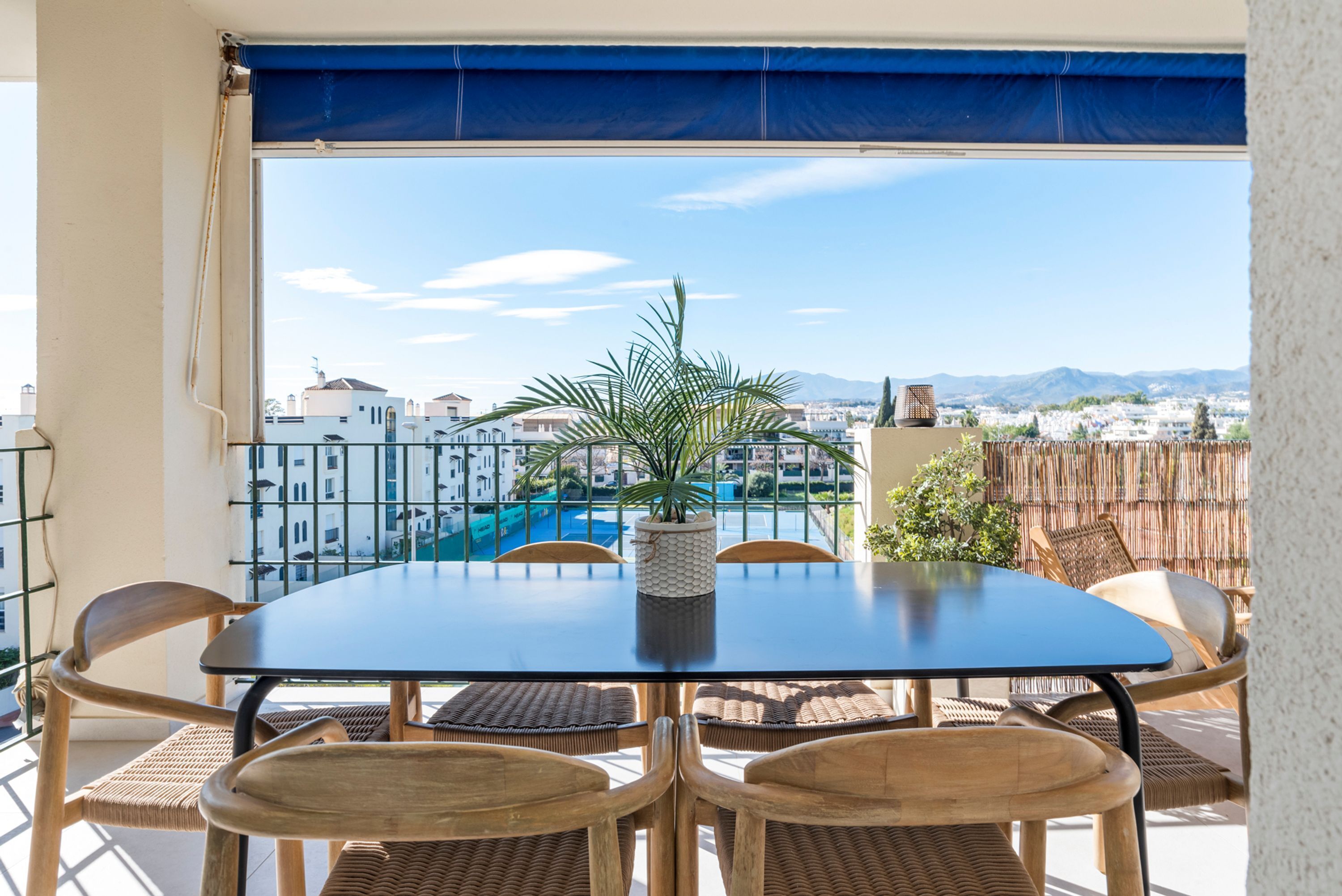 A balcony with a table and chairs, a potted plant, and a view of buildings and mountains.