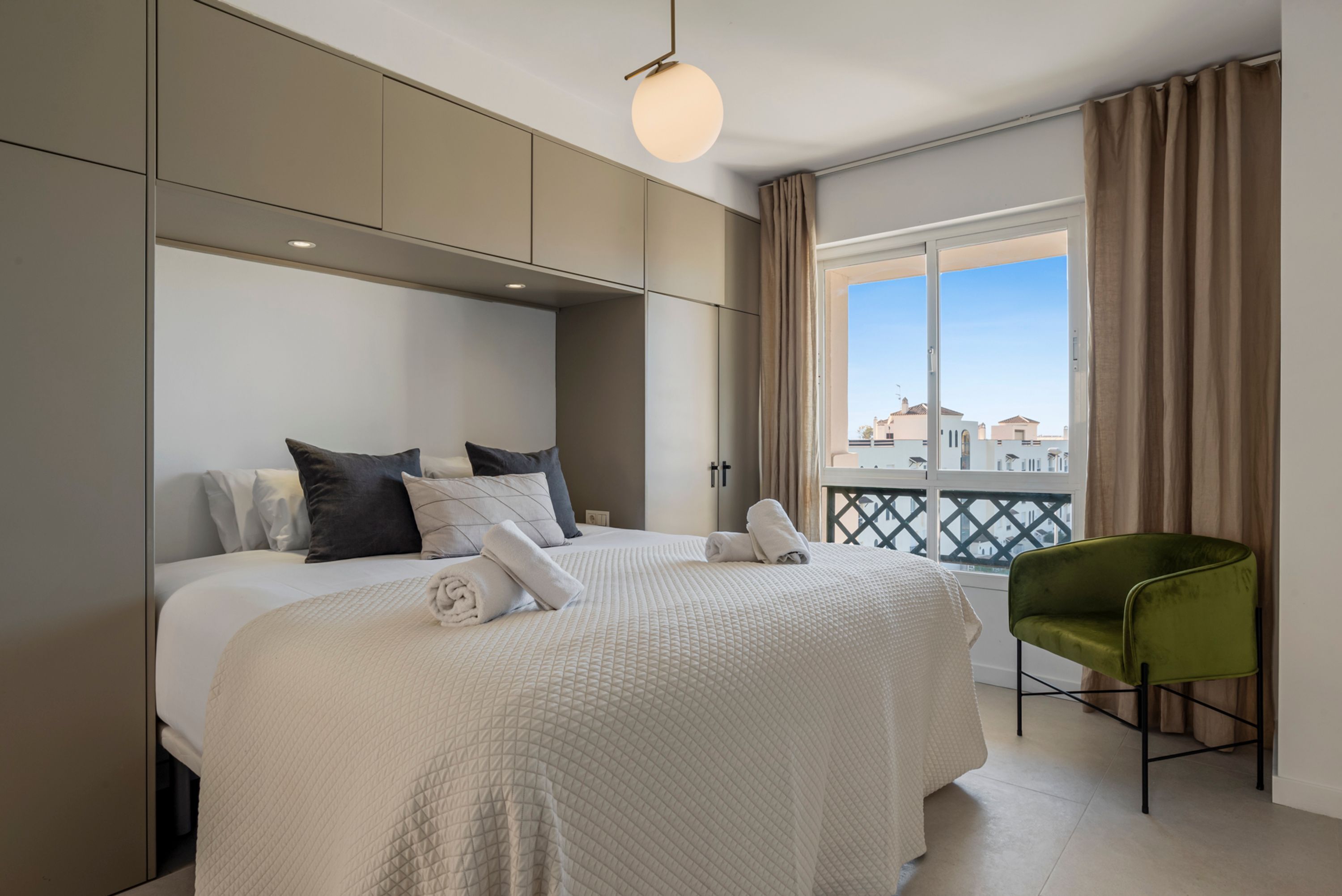 A hotel room with a large bed, a green chair, and a balcony with a view.