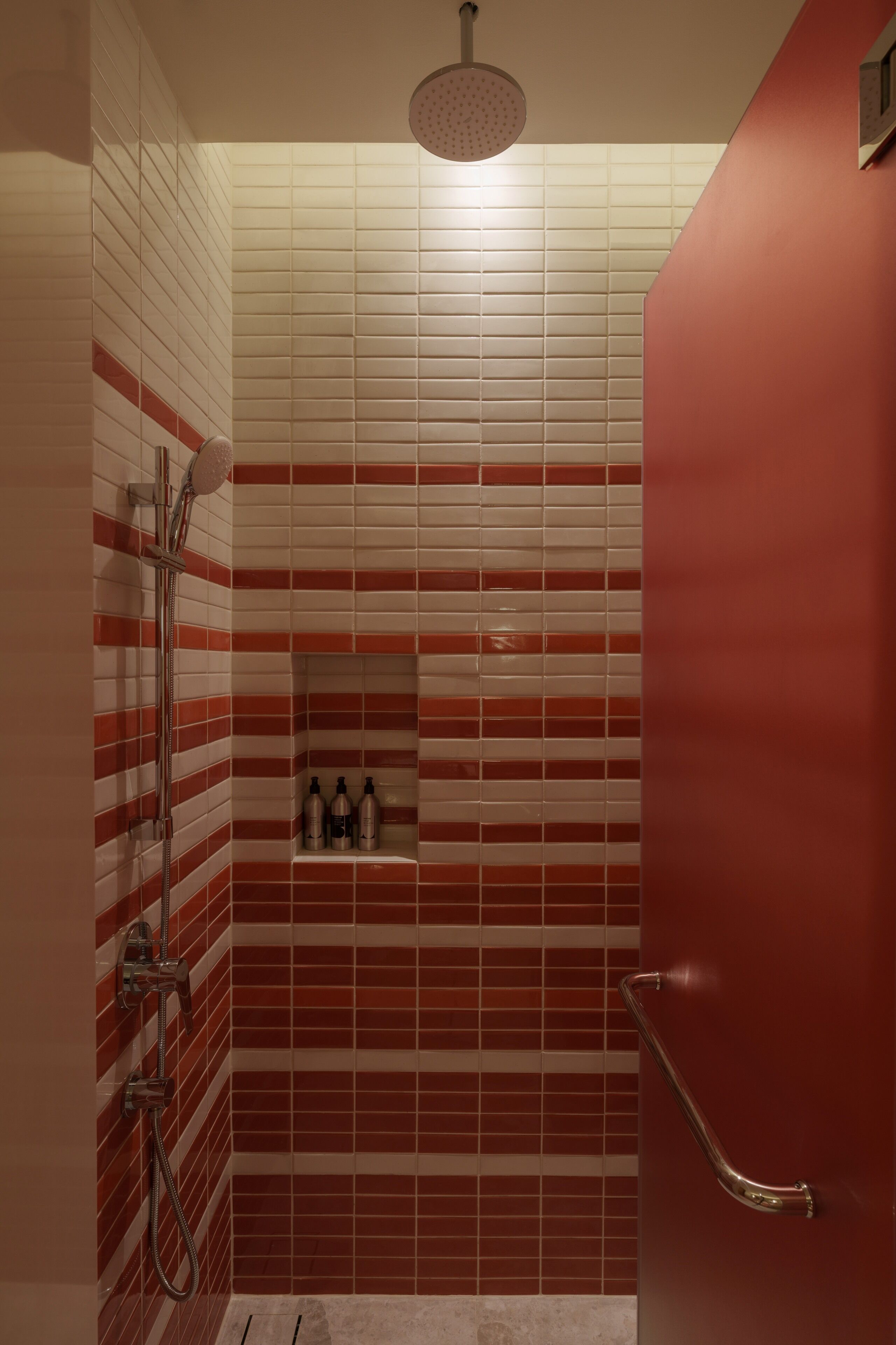 Eine Dusche mit roten und weißen Fliesen, ein Handbrausekopf und ein Ablagefach mit Toilettenartikeln.