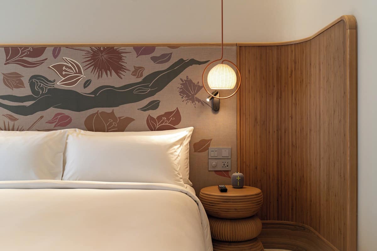 Una cama bien hecha con sábanas blancas, una lámpara de pared y una mesita de noche de madera con un control remoto.