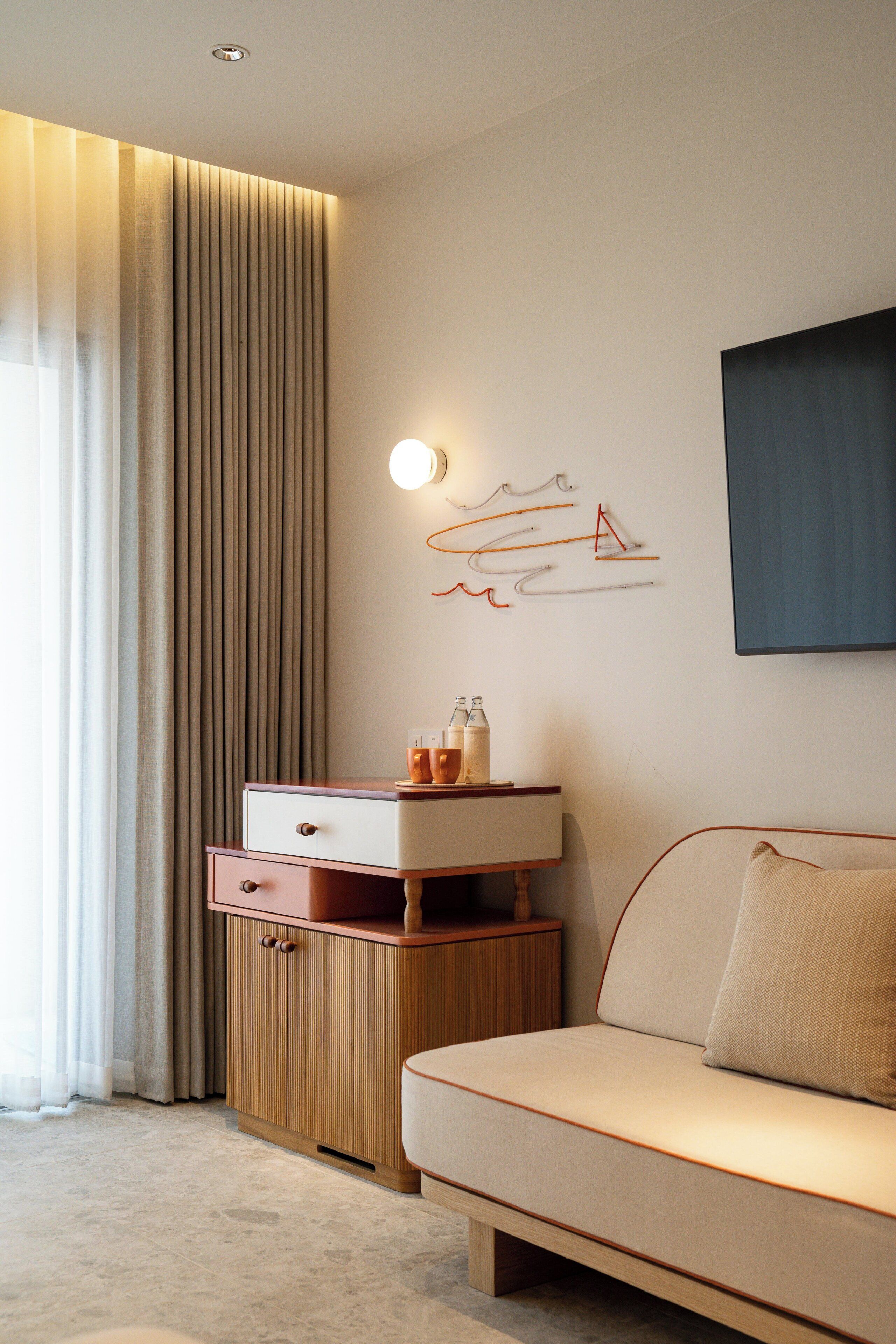 A modern hotel room with a sofa, a wooden cabinet, a wall-mounted light, and a large window with curtains.