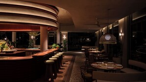 Un área de bar moderna con taburetes, una barra y luz ambiental. Hay mesas de comedor con sillas y un ventanal con vistas.