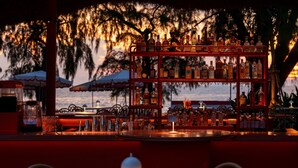 An outdoor bar area with a round table and chairs, a shelf of bottles, and umbrellas in the background. - The Standard Pattaya Na Jomtien (Sattahip)