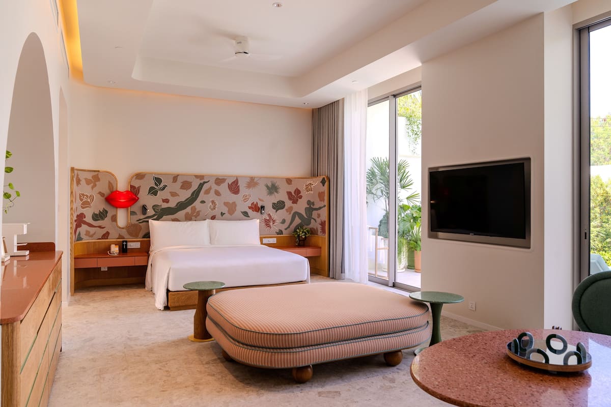 Habitación de hotel con cama, escritorio, silla, televisión y ventana con vista a follaje.