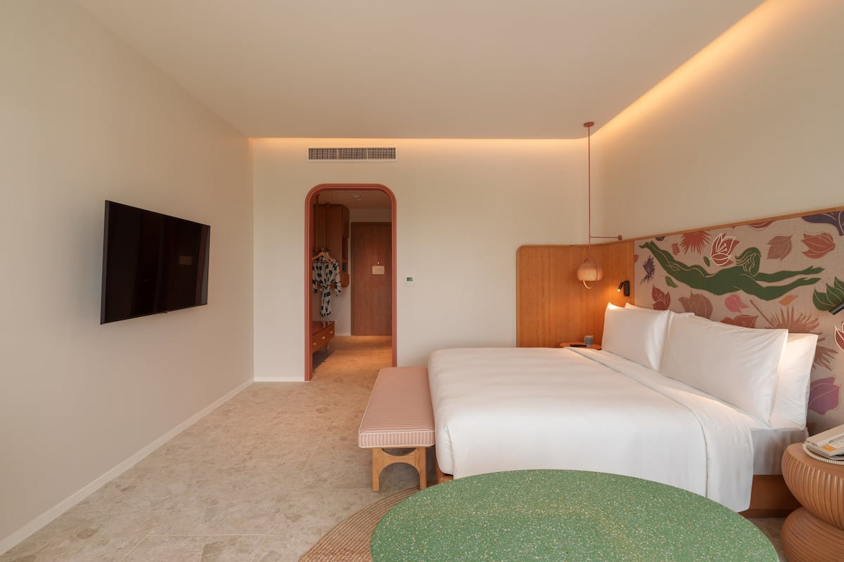 Habitación de hotel con cama, un banco, una mesa redonda, televisor montado en la pared y una puerta que da a un pasillo.