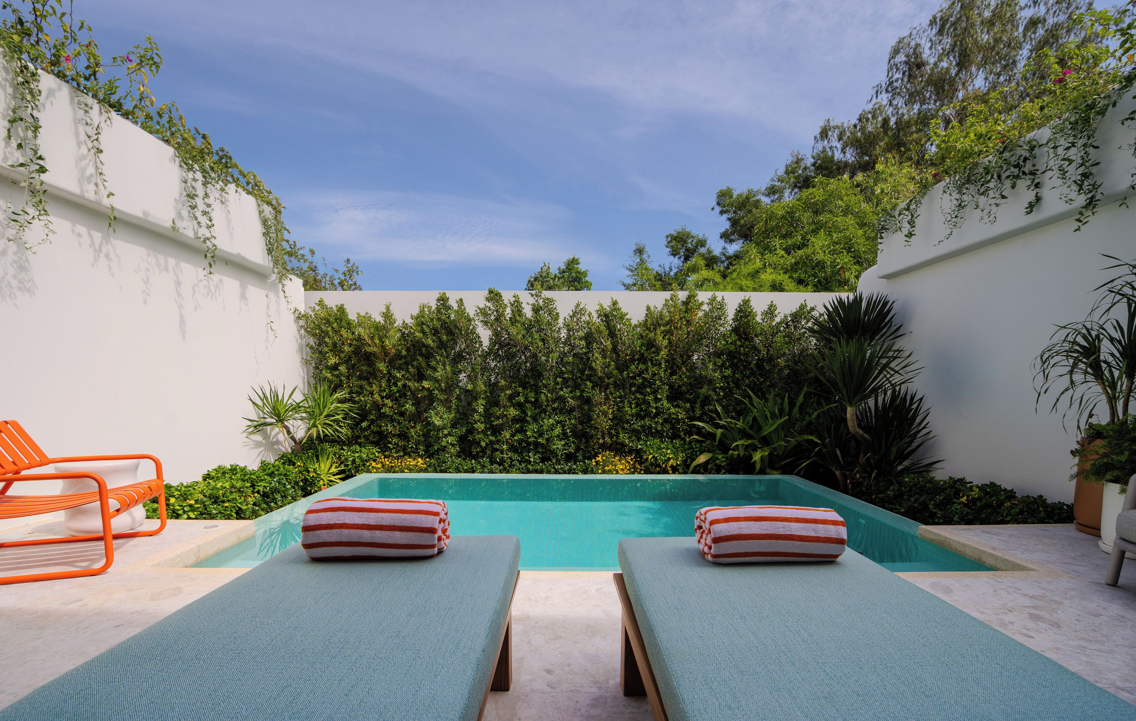 Zona de piscina con dos sillones de descanso y una piscina pequeña, rodeada de follaje y paredes blancas.